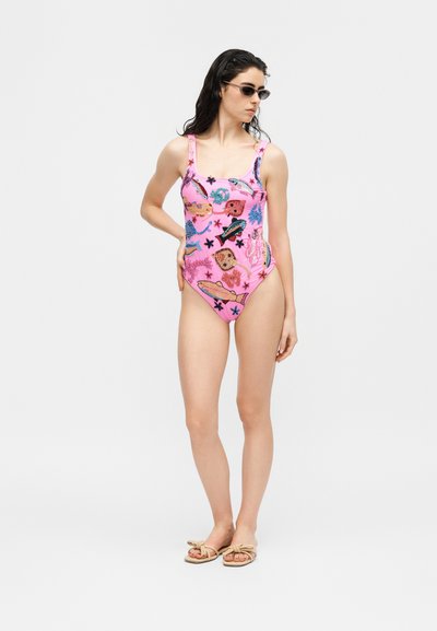 Γυναίκα που φοράει ένα ροζ ολόσωμο μαγιό με πολύχρωμα σχέδια θαλάσσιων ζώων, γυαλιά ηλίου και μπεζ σανδάλια, στέκεται μπροστά σε λευκό απλό φόντο.