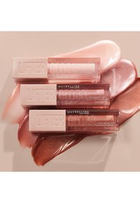 Trois tubes de gloss Lifter de Maybelline dans des teintes rose tendre et un emballage transparent, placés sur des échantillons brillants de teintes nude clair et de rose.