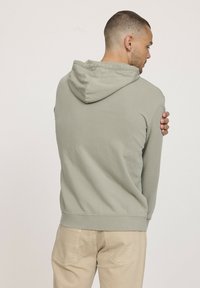 Sweatshirt à capuche vert clair avec une texture douce, des poignets et un ourlet côtelés, doté d'une poche kangourou, porté sur un pantalon beige.