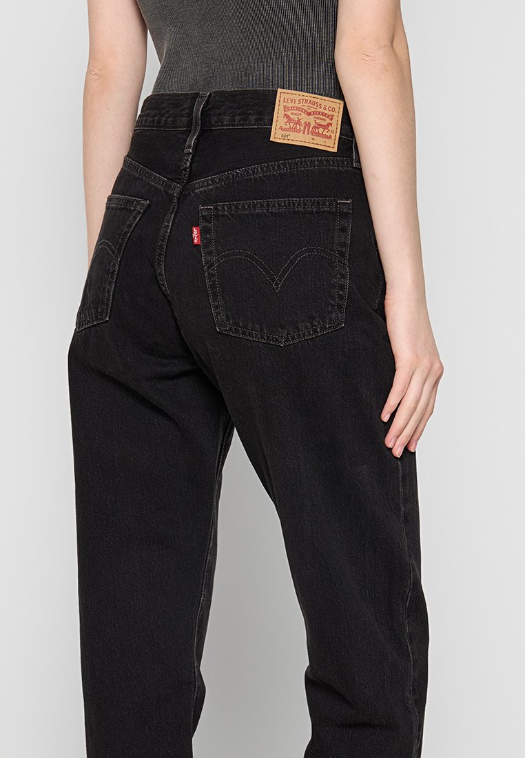 Jeans de mezclilla negra de cintura alta, con un parche de cuero en la parte trasera y costuras distintivas en los bolsillos traseros. Textura desgastada cerca de las costuras.
