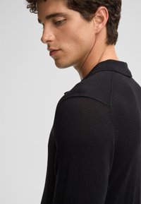 Polo shirt nera lavorata a maglia con finitura testurizzata, caratterizzata da un colletto e una vestibilità aderente. La vista laterale mostra i dettagli della cucitura della spalla.