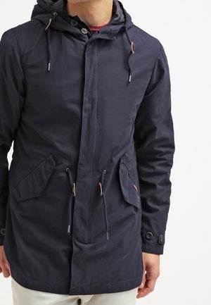 Homme portant une veste à capuche bleu marine avec poches avant, cordons à la capuche et à la taille, poignets boutonnés et fermeture éclair devant.