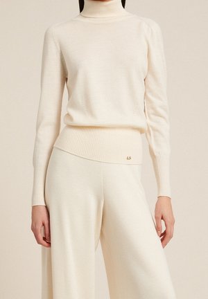 Femme portant un pull à col roulé en maille couleur crème et un pantalon large sur un fond beige uni.