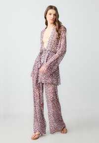Donna che indossa un abito leopardato rosa trasparente con pantaloni a gamba larga e una camicia a maniche lunghe con cintura, in piedi contro uno sfondo grigio chiaro uniforme.
