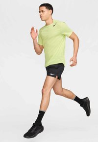 T-shirt de sport Nike vert clair pour homme, shorts noirs avec fentes latérales, chaussures noires et chaussettes noires avec accents gris, en position de course.