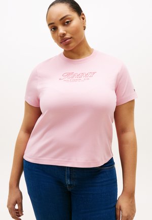 Femme portant un t-shirt rose clair Tommy Hilfiger et un jean bleu, debout devant un fond blanc uni.