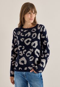 Marineblauer Pullover mit flauschiger Textur, der cremefarbene Leopardenflecken aufweist, einen runden Ausschnitt und lange Ärmel hat und mit hellblauen Jeans kombiniert ist.