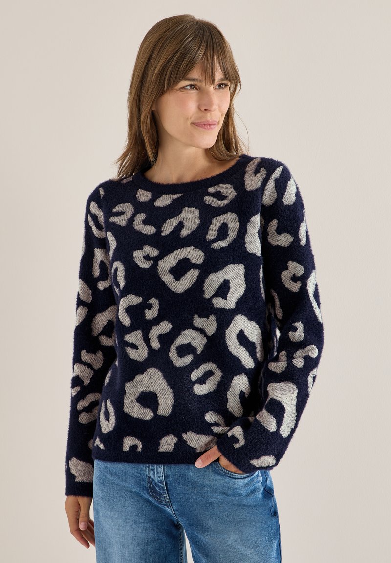 Marineblauer Pullover mit flauschiger Textur, der cremefarbene Leopardenflecken aufweist, einen runden Ausschnitt und lange Ärmel hat und mit hellblauen Jeans kombiniert ist.