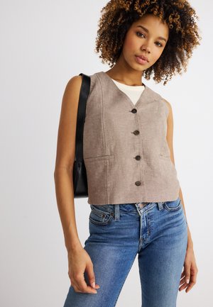 Young woman with curly hair wearing beige buttoned vest, white top, blue jeans, and black shoulder bag against plain light background.