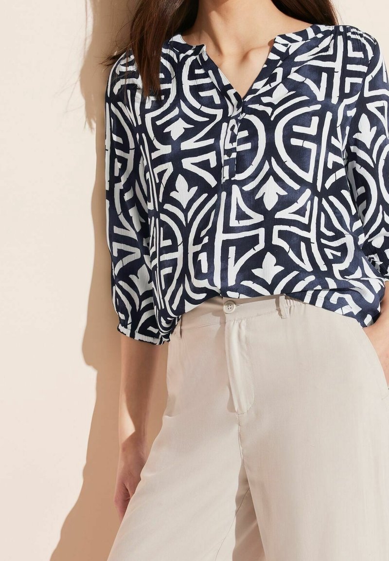 Blouse bleu marine avec motif géométrique blanc, manches 3/4, col en V et détail de boutons, assortie à un pantalon de couleur claire. Texture de tissu lisse.