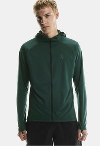 Junger Mann trägt eine dunkelgrüne Zip-Jacke mit Kapuze und Daumenlöchern und steht vor einem schlichten hellen Hintergrund.