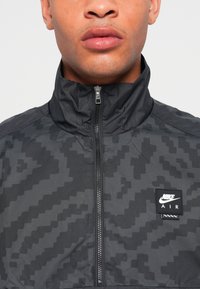 Nike AIR-jakke i svart med et subtilt pikselert mønster, høy krage, frontglidelås og logo-patch på venstre bryst.