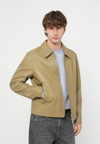 Δερμάτινο μπουφάν σε ανοιχτό μπεζ χρώμα με γιακά, φερμουάρ μπροστά και δύο πλαϊνές τσέπες. Φορεμένο πάνω από ένα γαλάζιο πουλόβερ με γκρι τζιν.