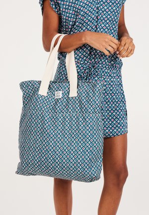 Tote bag met een blauw geometrisch patroon, voorzien van crèmekleurige stoffen handvatten, een geborduurd textuur en een logo-label op de voorkant.