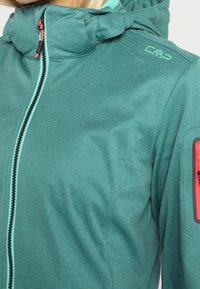 Veste softshell bleu sarcelle avec capuche, fermeture éclair frontale, poche zippée rouge sur la manche, et logo CMP sur l'épaule, portée par une personne.