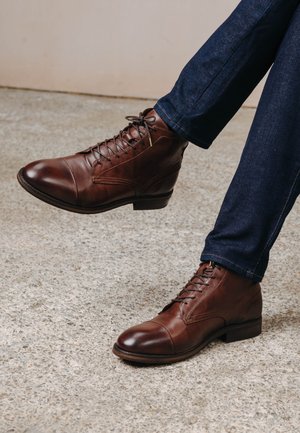 Brown leather lace-up ankle boots with a rounded toe, detailed stitching, and a low heel, paired with dark denim pants.
