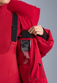 Piros szigetelt dzseki belső bal oldalán fényes okostelefon zsebbel, alatt pedig egy texturált kötött pulóver, állítható pántokkal.