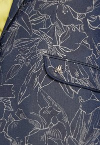 Tissu bleu foncé avec des contours floraux blancs, un petit rabat de poche, et une épingle décorative en métal argenté en forme de "M" attachée.