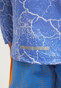 Chemise à manches longues bleue avec un motif blanc craquelé, ornée d'une bande argentée à l'ourlet. Associée à un short texturé orange et bleu.