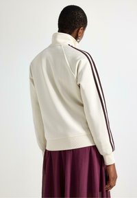 Krémová bunda s vysokým límcem, která má dvě tmavohnědé pruhy podél rukávů a vroubkované manžety, navržená pro volný střih.