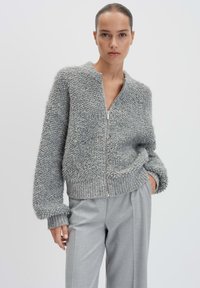 Cardigan grigio con zip realizzato in un tessuto fluffy e testurizzato. Presenta un colletto rotondo e polsini e orlo a costine, abbinato a pantaloni grigi chiari.