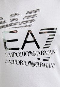 T-shirt bianca con loghi neri e argento, tra cui "EA7" e "EMPORIO ARMANI", con decorazioni in strass ed elementi grafici.
