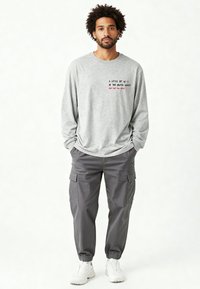 Uomo con capelli ricci che indossa una maglia a maniche lunghe grigio chiaro con scritta, pantaloni cargo grigio scuro, scarpe da ginnastica bianche, in piedi con le mani nelle tasche.