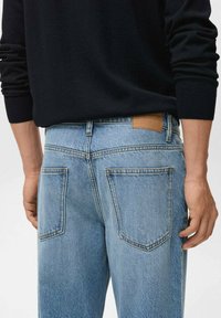 Una persona è in piedi indossando jeans azzurri chiari e un maglione nero, mostrando le tasche posteriori e la vita con un'etichetta in pelle beige.