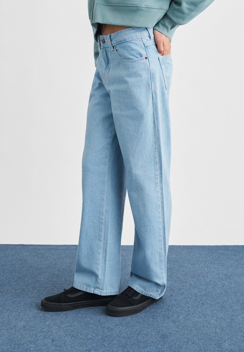 Persoon gekleed in lichtblauwe wijde jeans en zwarte sneakers, staand op een blauw tapijt met een hand in de achterzak.