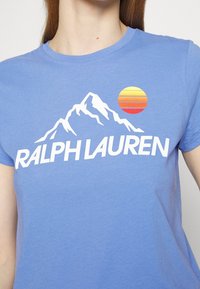 Modré bavlněné tričko s bílým textem "RALPH LAUREN," grafikou hory a kruhovým designem západu slunce v žluté, oranžové a červené.