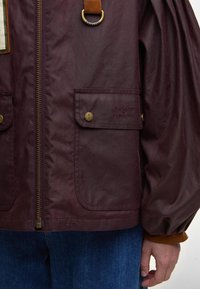 Μπορντώ αδιάβροχη ζακέτα με φερμουάρ, δύο μπροστινές τσέπες, χρυσές λεπτομέρειες, μπαλόνι μανίκια και καφέ δερμάτινη λεπτομέρεια στον ώμο.