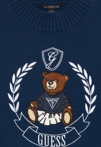 Mornarska pulover s plišasto vezenino medveda, ki nosi modro majico in belo krilo, obdanega z belimi lovorjevimi listi in logotipom.