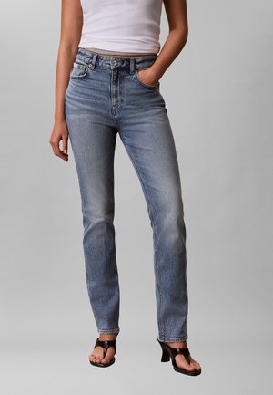 Woman wearing high-waist faded blue jeans, white sleeveless top, black heeled sandals, with one hand in pocket, standing against plain background.