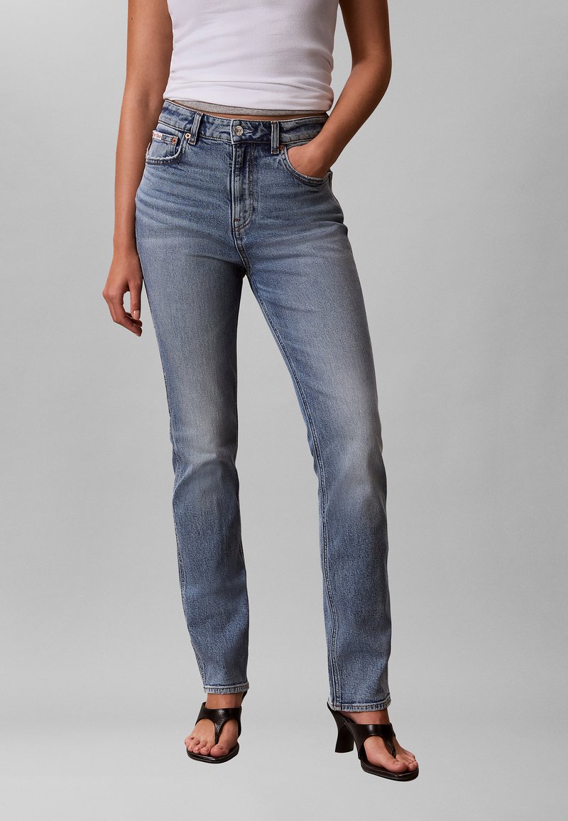 Nő magas derekú, halványkék farmerben, fehér ujjatlan felsőben, fekete magassarkú szandálban, egy kézzel a zsebében áll, egyszínű háttér előtt.