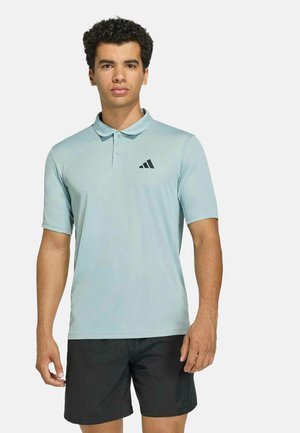 Joven de pie con una camiseta polo de manga corta azul claro con el logo de Adidas y pantalones cortos negros sobre un fondo liso.