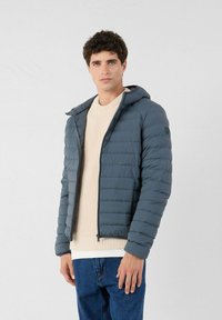 Gevoerde jas in donkergrijs met een capuchon, met horizontale gestikte textuur en een rits aan de voorkant. Draag het over een beige gebreide trui en jeans.