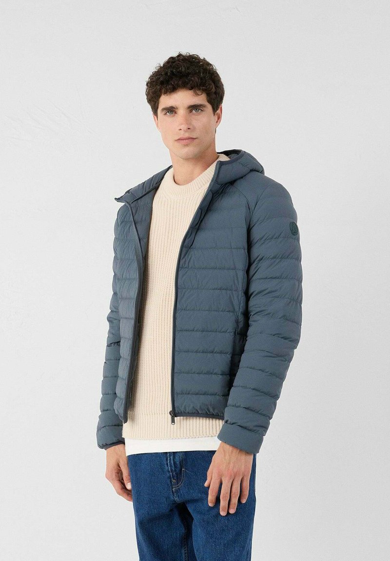 Gevoerde jas in donkergrijs met een capuchon, met horizontale gestikte textuur en een rits aan de voorkant. Draag het over een beige gebreide trui en jeans.