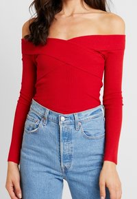 Body rosso a costine, senza spalle e con maniche lunghe. Abbinato a jeans a vita alta di colore blu chiaro, con tasche e chiusura a bottone.