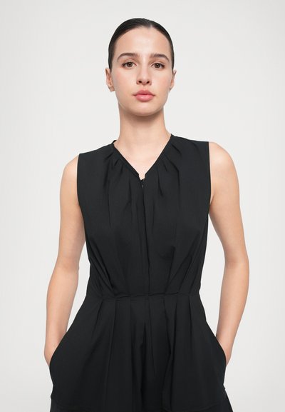 Vestido negro sin mangas con escote en V, detalles plisados y bolsillos laterales. La tela tiene una textura suave y una silueta fluida.