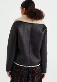 Svart läderjacka med en krämfärgad fuskpälskrage, som har en rak skärning och synliga sömnadsdetaljer på baksidan.