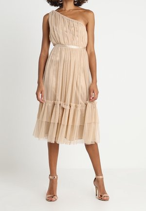 Kvinde iført en beige, asymmetrisk midikjole med plissé og flæsekant samt matchende højhælede sandaler, stående foran en ensfarvet baggrund.