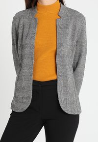 Una donna sta in piedi indossando un blazer a quadri grigio su un dolcevita a coste arancione brillante, abbinato a pantaloni neri, su uno sfondo semplice.