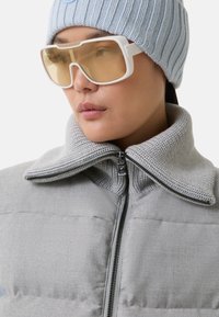 Lys grå polstret jakke med en strikket krave, lynlåslukning og to forlommer. Sammen med hvide oversized solbriller og en blå strikket hue.