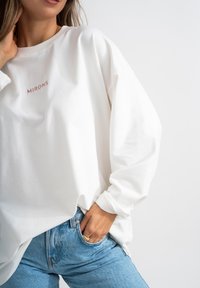 Maglione bianco a maniche lunghe, con logo "MIRONS" ricamato in rosa. Indossato con jeans a vita alta blu chiaro. Texture liscia.