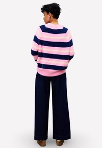 Personne avec des cheveux bouclés courts portant un pull rayé rose, bleu marine et crème, un pantalon ample sombre et des chaussures marron, de dos.