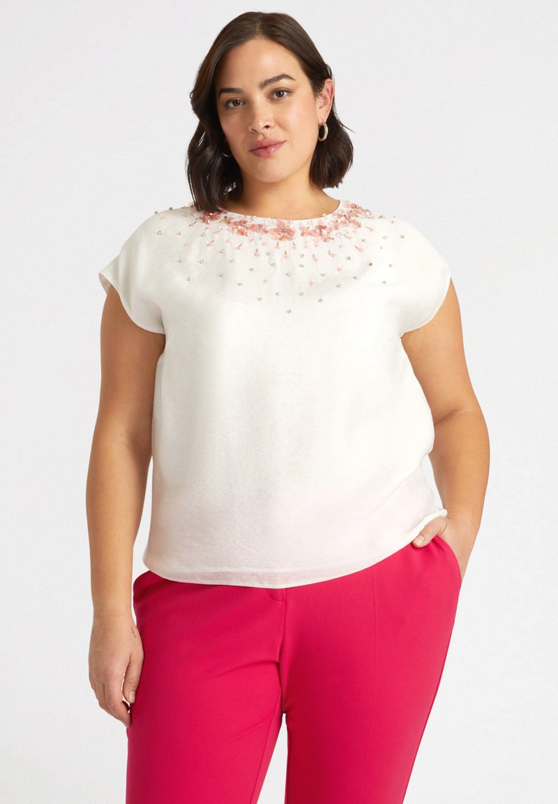 Blusa bianca a maniche corte con ricamo di perline floreali sul colletto. Presenta una vestibilità rilassata e una texture morbida. Abbinata a pantaloni rosa.