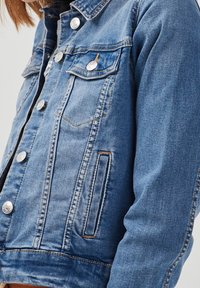 Veste en denim bleu clair avec col classique, boutons argentés, deux poches poitrine et détails de couture à l'avant. Texture douce avec coupe décontractée.