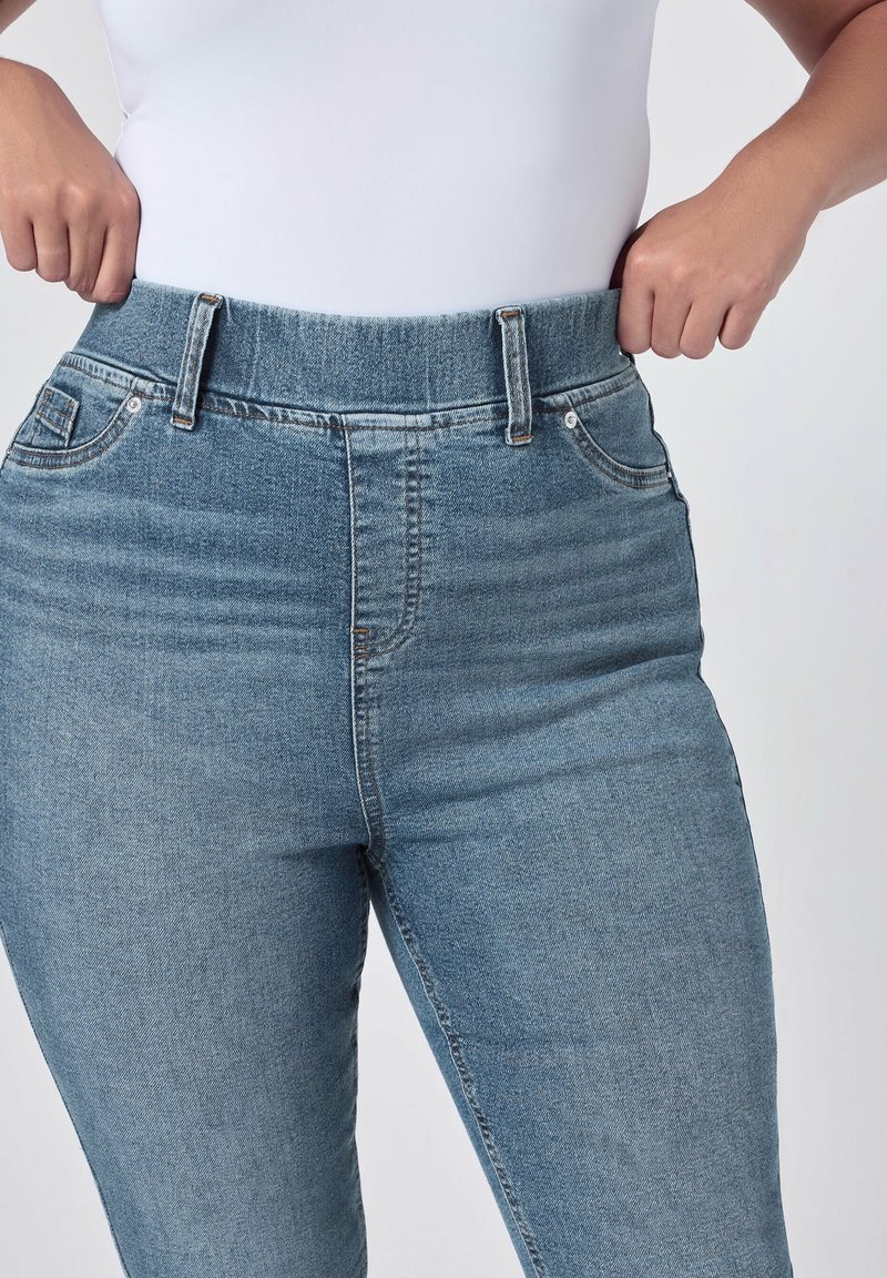 Personne portant un jean bleu et un haut blanc tire la taille du jean vers l'extérieur pour montrer la coupe ou l'élasticité.