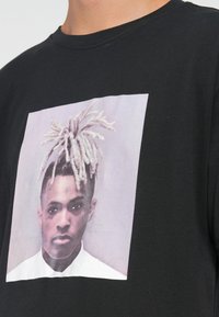 Sort bomulds T-shirt med et stort grafisk print af en person med lyse dreadlocks og et neutralt udtryk på en farvet baggrund.