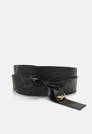 Large ceinture en cuir noir nouée en nœud avec un petit détail métallique doré en forme d’oiseau à une extrémité sur un fond uni.